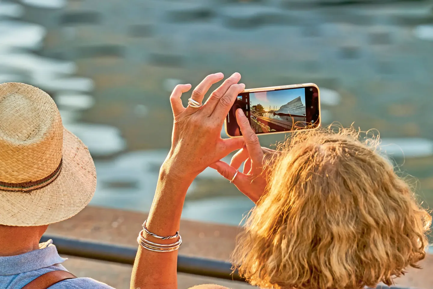 © Bregenzer Festspiele / Anja Koehler Frau mit lockigem Haar hält Smartphone hoch und macht Foto von Wasser und Gebäuden neben einem Mann mit Strohhut.
