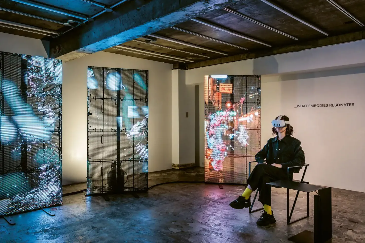 Person mit VR-Brille sitzt auf Stuhl in dunklem Raum mit großflächigen, abstrakten digitalen Projektionen auf Wänden und Trennwänden