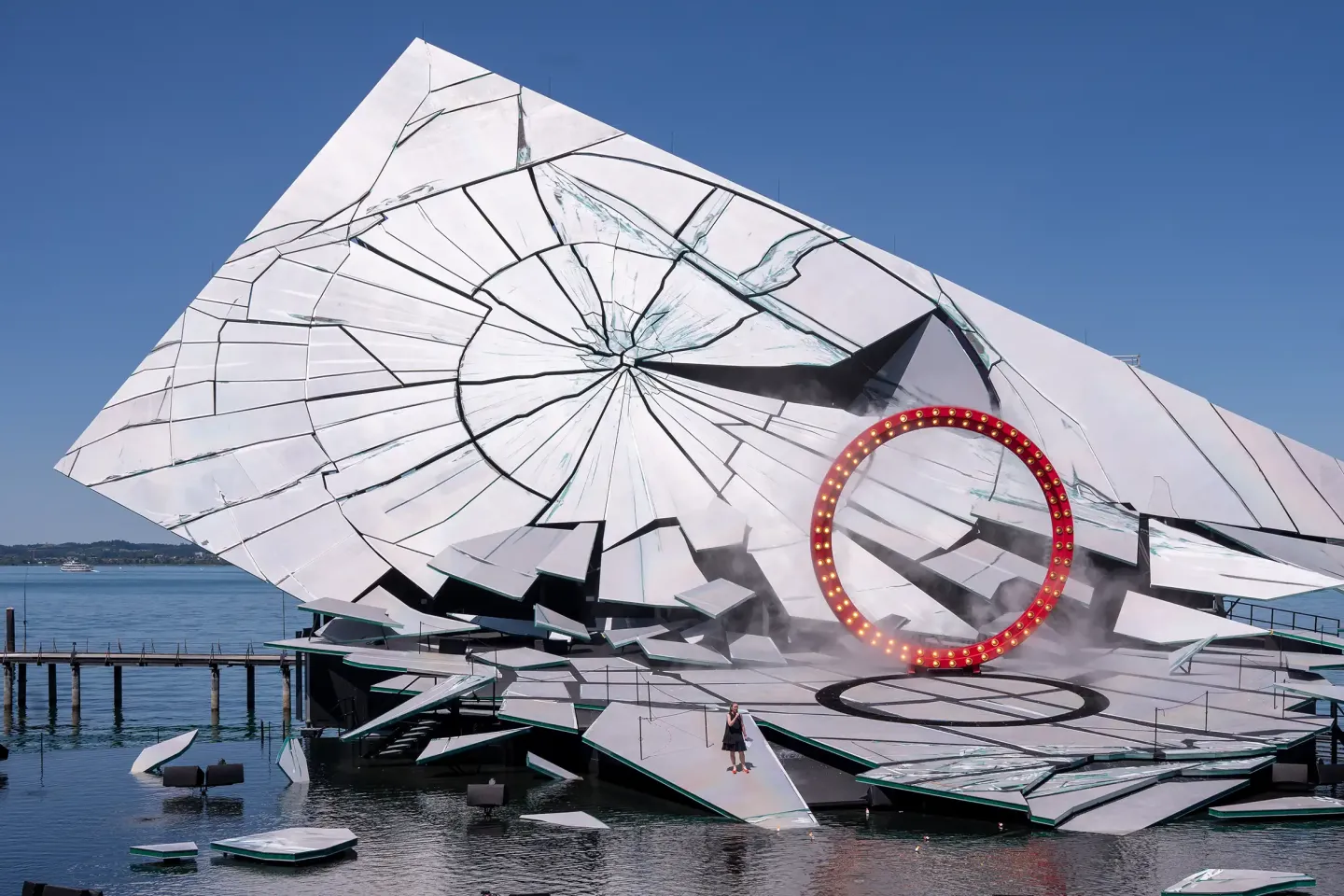 © Bregenzer Festspiele / Christian Lins Große, schräg stehende Bühnenkonstruktion mit weiß-schwarzer Risszeichnung und rotem, beleuchtetem Ring auf einem See mit Steg und mehreren kleinen Booten.