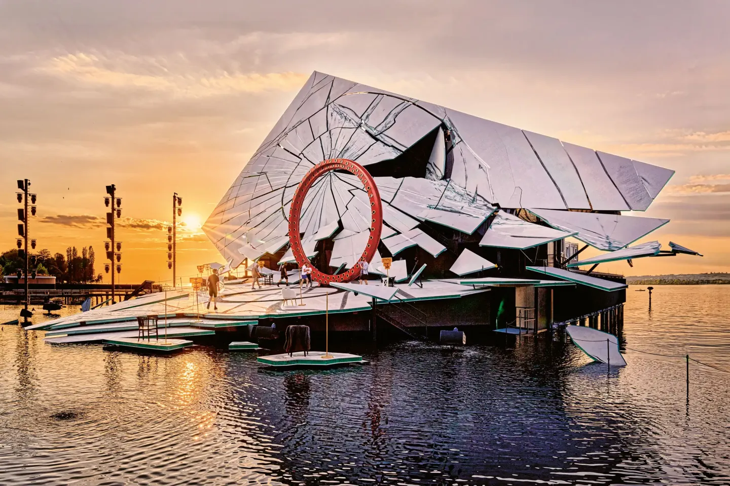 © Bregenzer Festspiele / Anja Koehler Seebühne mit großem roten Ring und spiegelnder, geometrisch gebrochener Fassade bei Sonnenuntergang auf einem See.