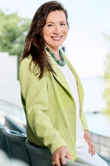 Lilli Paasikivi, Intendantin der Bregenzer Festspiele, lächelnd im Freien mit grüner Jacke und weißem Outfit vor verschwommenem Hintergrund mit Bäumen und Wasser