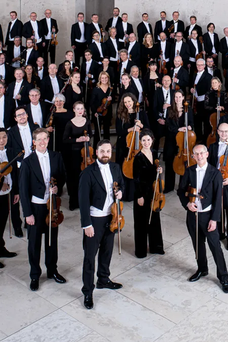 Mitglieder der Wiener Symphoniker in formeller Konzertkleidung mit Instrumenten, auf einer Treppe in einem modernen Saal gruppiert