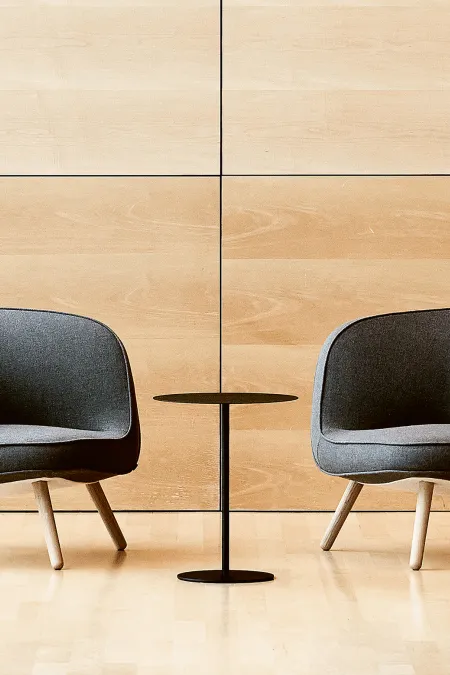 Zwei graue Sessel mit Holzbeinen und ein kleiner runder schwarzer Tisch vor einer hellen Holzpaneelwand