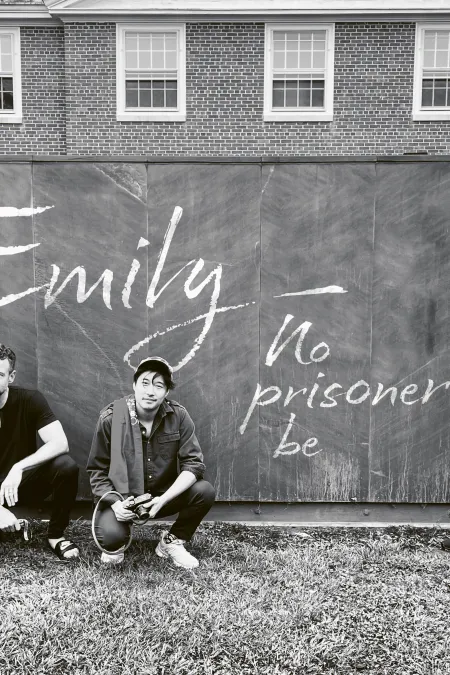 Fünf Personen vor einer Tafel mit der Aufschrift 'Emily - no prisoner be', Kevin Puts, das musikalische Trio time for Three und Joyce DiDonato, entspannt im Freien vor einem Backsteingebäude