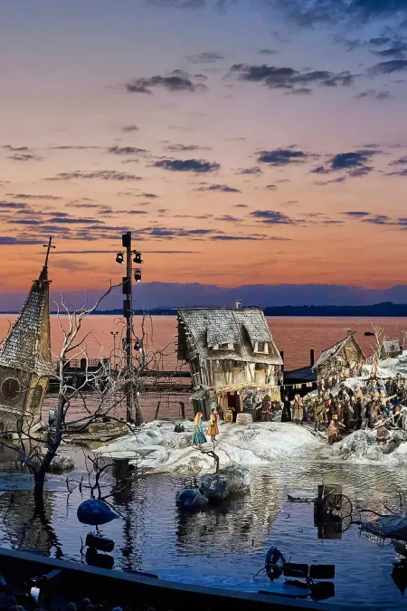 Kulisse "Der Freischütz" - Schneelandschaft überflutet, windschiefe Häuser, ein Kirchturm ragt aus dem Wasser, dramatischer Sonnenuntergang im Hintergrund in orange bis blau