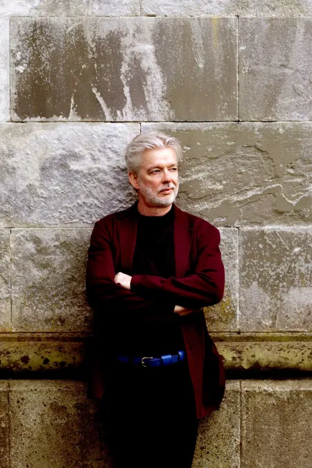 © Felix Broede Dirigent Jukka Pekka Saraste lehnt an einer Mauer, rotes Sakko, dunkle Hose