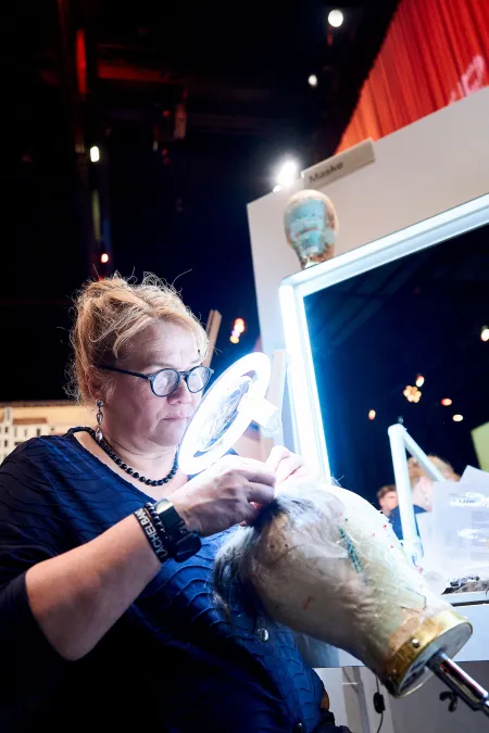 Chefmaskenbilnerin Frauke Gose mit Ringlicht am Perückenknüpfen
