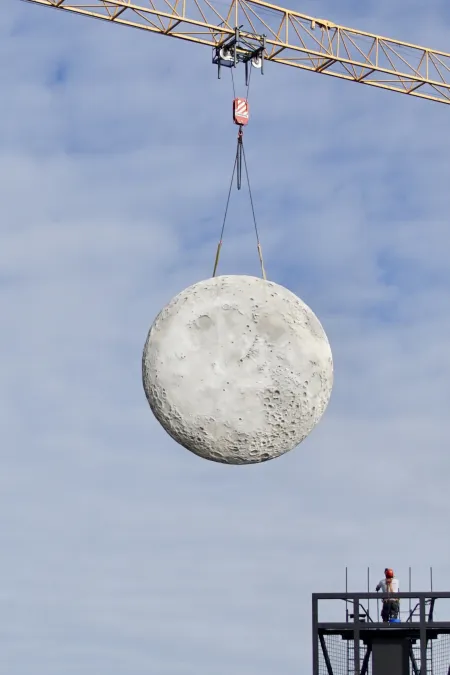 Runder, kraterübersäter Mond wird von einem Baukran an zwei Seilen über einem Gerüst mit einer Person gehoben.