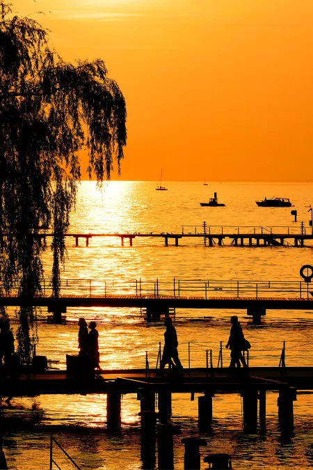 Silhouetten von Menschen auf Stegen vor einem See bei Sonnenuntergang mit orangefarbenem Himmel und mehreren Booten auf dem Wasser.