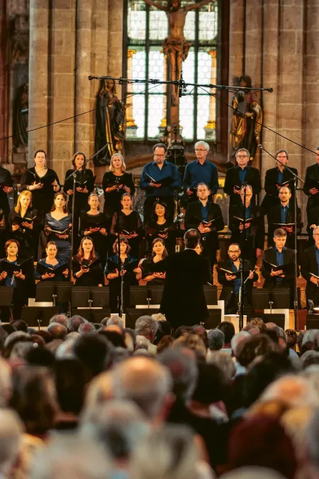 Großer Chor mit mehreren Sängerinnen und Sängern in dunkler Kleidung steht auf Stufen in einer Kirche vor Publikum, Dirigent mit erhobenen Armen.