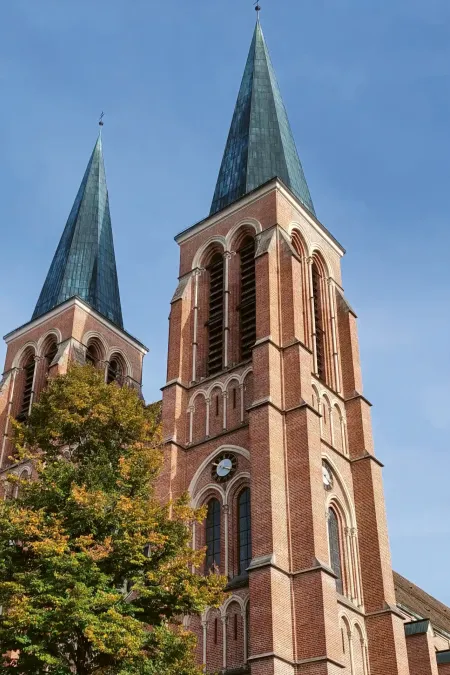 Zwei hohe, spitz zulaufende Kirchtürme aus rotem Backstein mit Bäumen im Vordergrund und blauem Himmel.