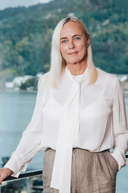 Frau mit langen blonden Haaren trägt weiße Bluse mit Schluppe und graue Hose, steht an Geländer mit Wasser und bewaldetem Hügel im Hintergrund.