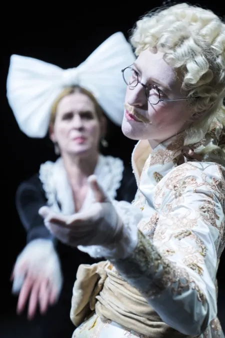 Zwei Personen in historischen Kostümen, eine mit weißer Perücke und besticktem Jackett, die andere mit großem weißem Haarschmuck, beide strecken die Hände aus.