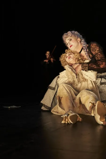 Zwei Personen in historischen Kostümen, eine sitzt am Boden, die andere hält sie im Arm, im Hintergrund eine Person mit Geige.