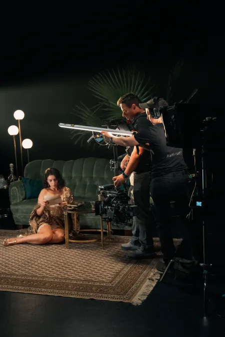 © Bregenzer Festspiele / Jonatan Langendorf Frau in goldfarbenem Kleid sitzt auf Teppich vor grünem Sofa, zwei Männer bedienen Filmkamera und Lichttechnik.