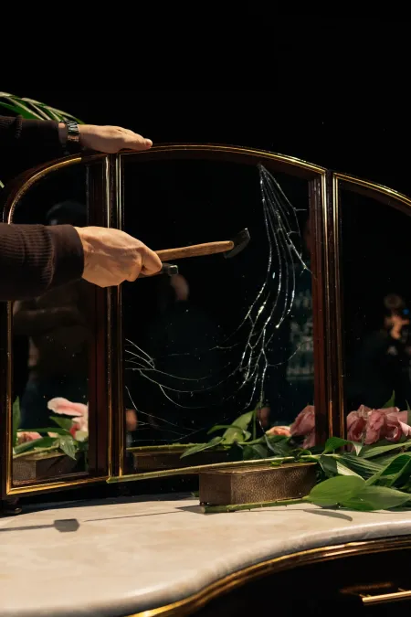 © Bregenzer Festspiele / Jonatan Langendorf Hände halten Hammer und zerbrechen einen dreiteiligen Spiegel mit goldfarbenem Rahmen auf einer Marmorplatte, umgeben von grünen Blättern und rosa Blumen.