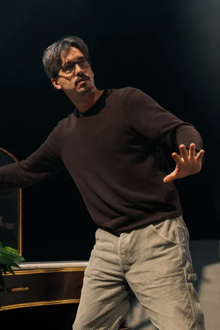 Mann in schwarzem Pullover und hellen Hosen steht mit ausgestreckter Hand vor einem Spiegel mit Blumen auf dem Tisch.