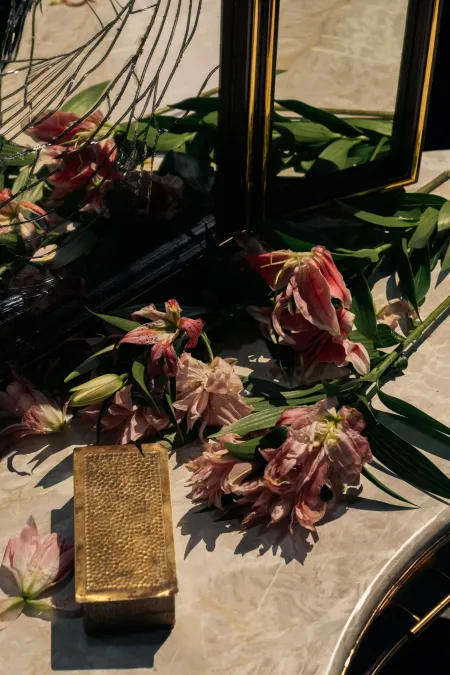 © Bregenzer Festspiele / Jonatan Langendorf Verwelkte rosa Lilien und grüne Blätter liegen neben einem goldfarbenen, rechteckigen Schmuckkästchen auf einer Marmoroberfläche.
