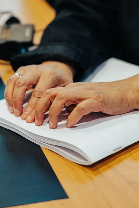 Hände ertasten ein aufgeschlagenes Braillebuch auf einem Tisch