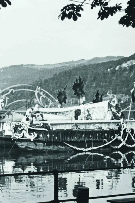 Black-and-white photo of a boat decorated with ornaments on a lake, with forested hills in the background and a tree in the foreground.