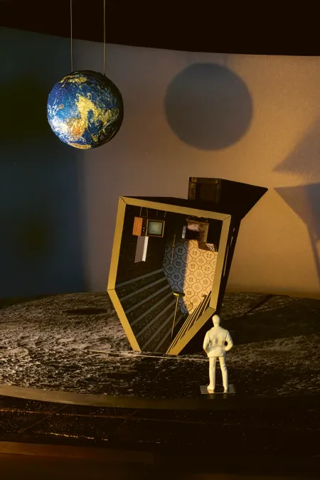 Miniaturfigur steht vor einem schräg aufgestellten, offenen Modell eines Raumes mit Treppe und Fernseher unter einer hängenden Erdkugel