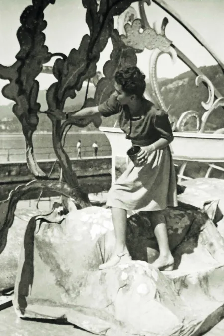 A woman in a skirt and sweater is standing barefoot on a large, irregular sculpture, holding a small bucket in her hand.
