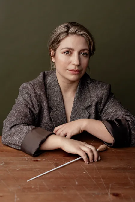 Person mit grauem Blazer sitzt an einem Holztisch und hält einen Dirigentenstab