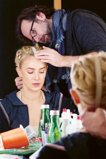 © Bregenzer Festspiele / Anja Koehler A person is measuring the head of a seated person with a tape measure in front of a mirror.