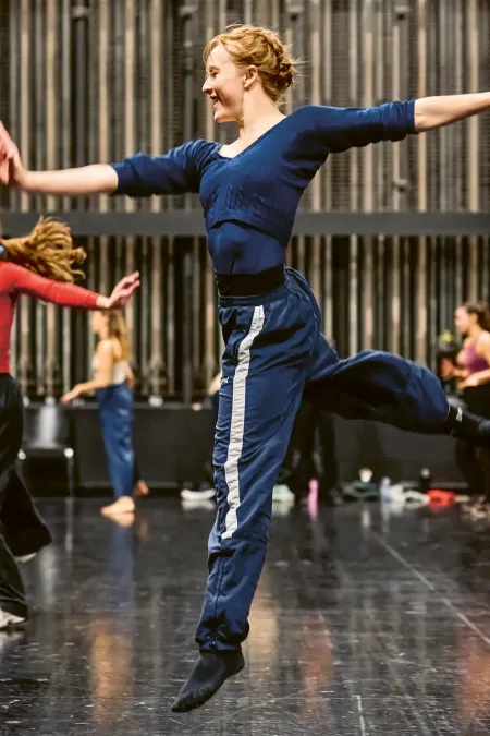 © Bregenzer Festspiele / Anja Koehler Tänzerin in blauer Trainingskleidung springt mit ausgestreckten Armen und einem angewinkelten Bein auf einer Bühne mit weiteren tanzenden Personen im Hintergrund