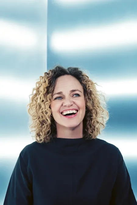 Anna Kelo © Veikko Kähkönen Person mit lockigem Haar und schwarzem Oberteil vor hell gestreifter Wand
