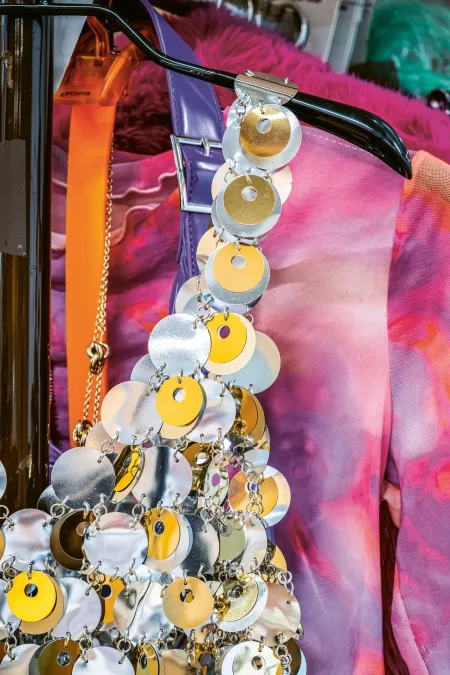 Kostüm mit großen, runden, silbernen und goldfarbenen Pailletten an einem schwarzen Träger vor einem pink-violett gemusterten Kleid auf einem Kleiderbügel.