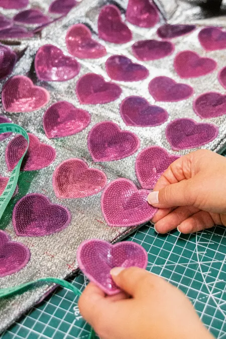 Hände legen pinke Herzapplikationen auf silberglänzenden Stoff mit Maßband auf grünem Schneideuntergrund.