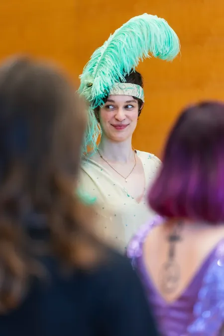 © Bregenzer Festspiele / Kristina Becker Frau mit hellgrünem Federkopfschmuck und hellgrünem Kostüm lächelt zwischen zwei unscharfen Personen