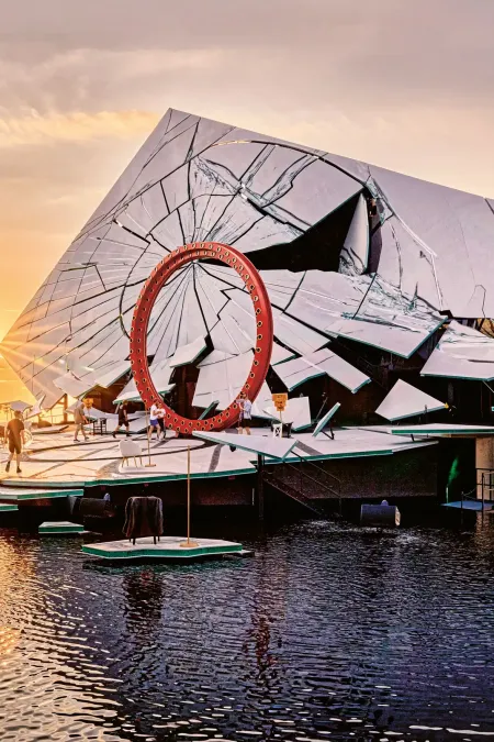 Seebühne mit großem roten Ring und spiegelnder, geometrisch gebrochener Fassade bei Sonnenuntergang auf einem See.