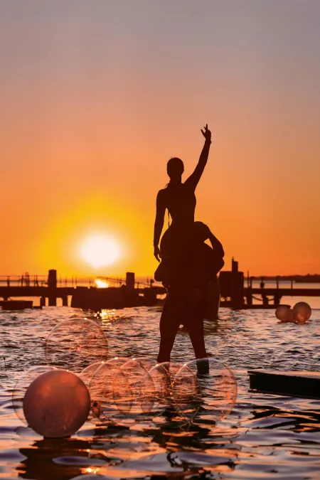 © Bregenzer Festspiele / Anja Koehler Silhouetten von zwei Personen im Wasser bei Sonnenuntergang, eine Person trägt die andere auf den Schultern, im Vordergrund schwimmende Kugeln und im Hintergrund ein Steg.