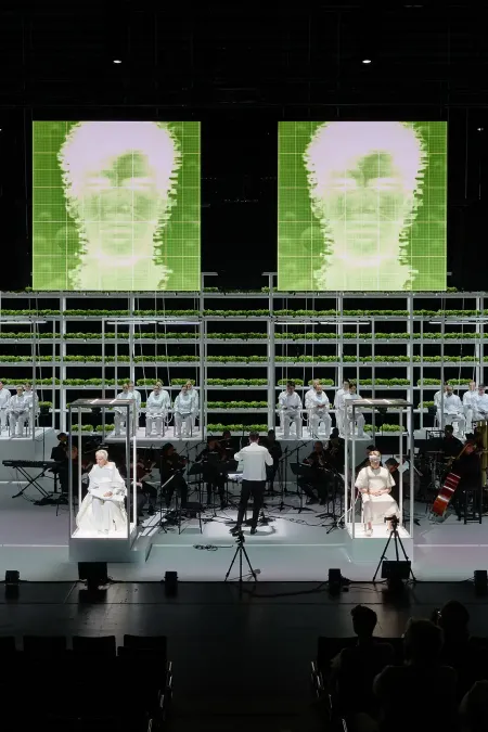 Bühnenbild mit mehreren Musikern und Sängern in weißen Kostümen vor einer Wand aus Pflanzenkästen und vier großen, grünen, digital dargestellten Gesichtern