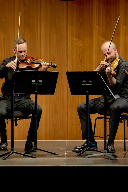Vier Musiker sitzen auf Stühlen vor Holzpaneelen und spielen Streichinstrumente, zwei mit Violinen, einer mit Bratsche, einer mit Cello.