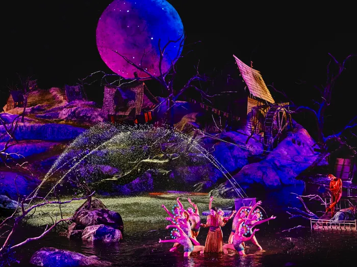 Bühnenbild von 'Der Freischütz' mit Tänzern in farbenfrohen Kostümen vor einem großen, leuchtenden Vollmond und einer mystischen Waldkulisse