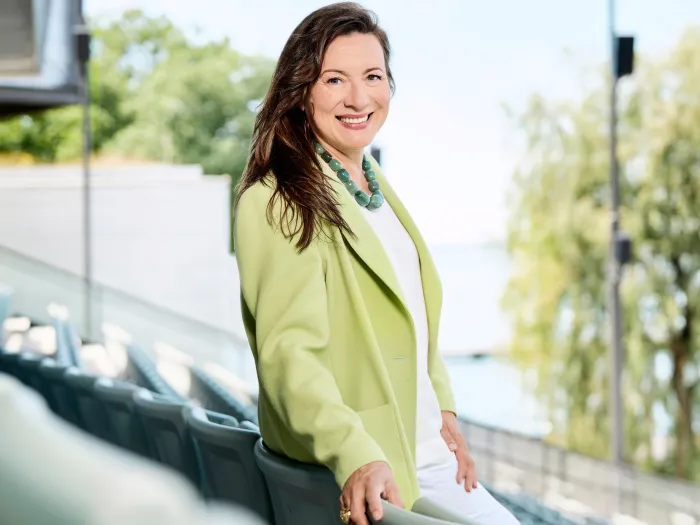 Lilli Paasikivi, Intendantin der Bregenzer Festspiele, lächelnd im Freien mit grüner Jacke und weißem Outfit vor verschwommenem Hintergrund mit Bäumen und Wasser