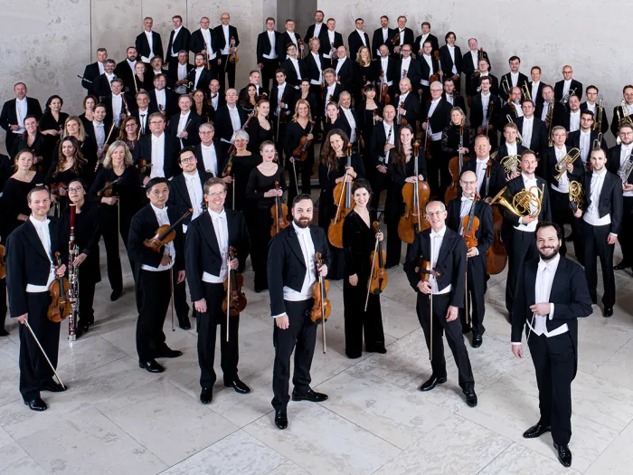 Mitglieder der Wiener Symphoniker in formeller Konzertkleidung mit Instrumenten, auf einer Treppe in einem modernen Saal gruppiert