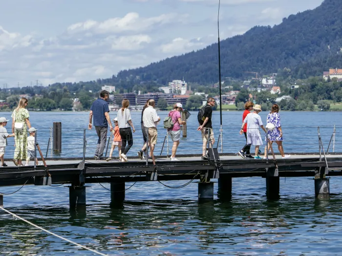 Gruppe von Menschen, darunter Kinder und Erwachsene, die auf einem Steg über einem See spazieren, im Hintergrund bewaldete Hügel und Häuser