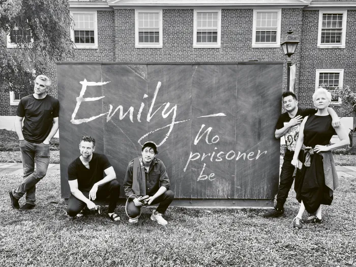 Fünf Personen vor einer Tafel mit der Aufschrift 'Emily - no prisoner be', Kevin Puts, das musikalische Trio time for Three und Joyce DiDonato, entspannt im Freien vor einem Backsteingebäude