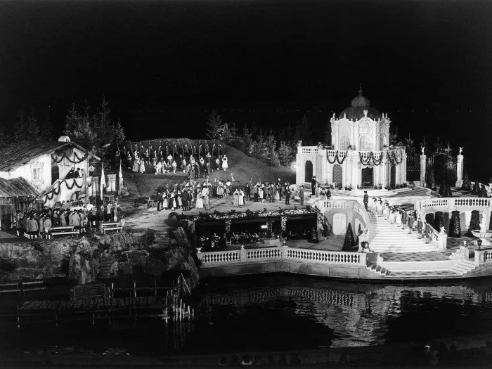 © Bregenzer Festspiele Szene aus "Der Vogelhändler", 1984 auf der Seebühne, zweistöckiges Bühnenbild, auf der oberen Ebene links befindet sich ein Gebäude und rechts ein zweistöckiger Pavillon, vor dem Pavillon führt eine Treppe in die untere Ebene, in der Mitte der Bühne sind viele kostümierte Darstellende
