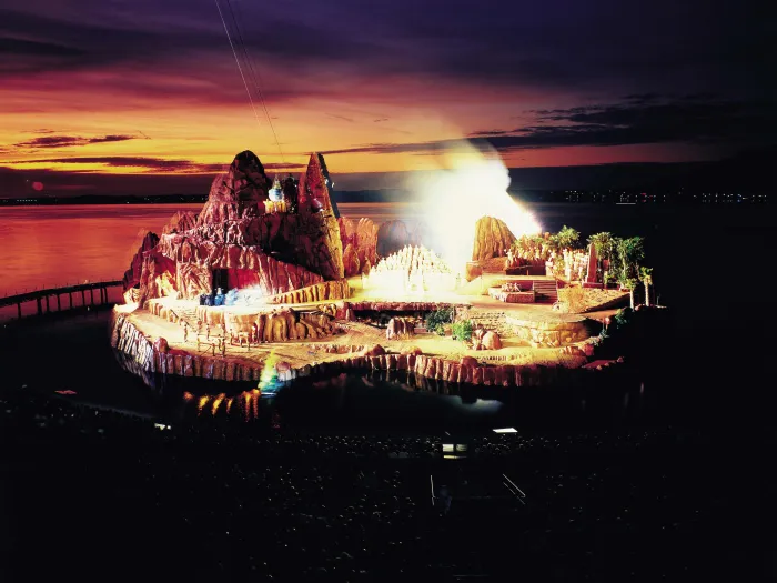 Szene aus "Die Zauberflöte", 1986 auf der Seebühne, Felsenlandschaft mit einzelnen Bäumen, ein größerer Berg befindet sich auf der linken Seite des Bühnenbilds, die Darstellenden sind mittig aufgereiht und hell angeleuchtet 