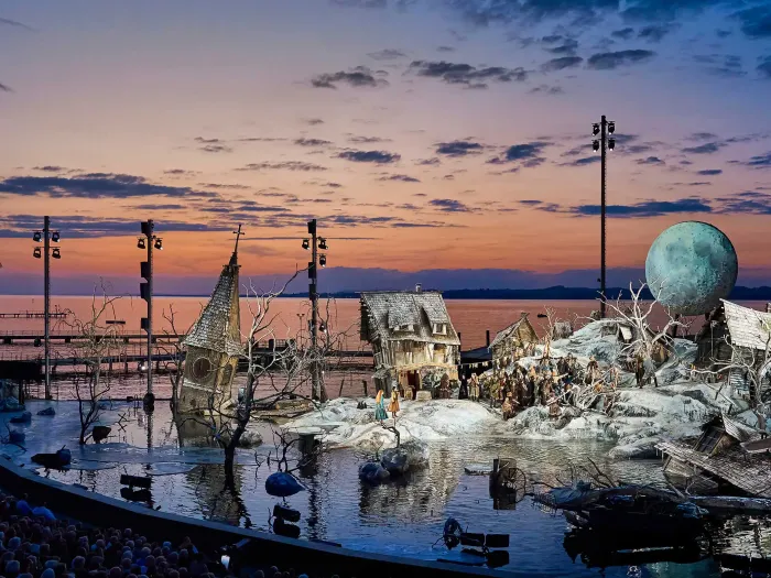 © Bregenzer Festspiele / Anja Köhler Kulisse "Der Freischütz" - Schneelandschaft überflutet, windschiefe Häuser, ein Kirchturm ragt aus dem Wasser, dramatischer Sonnenuntergang im Hintergrund in orange bis blau