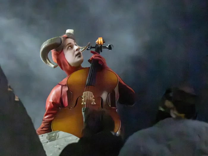 Person mit Hörnern auf dem Kopf spielt Cello auf einer Bühne mit dunklem Hintergrund