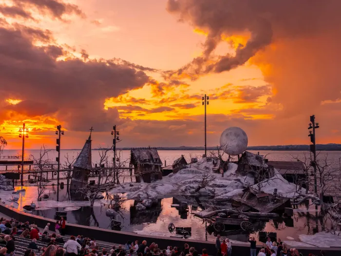 Freilichtbühne mit winterlicher Kulisse und großem Vollmond-Requisit vor einem dramatischen Sonnenuntergang am See, Publikum sitzt auf Tribünen