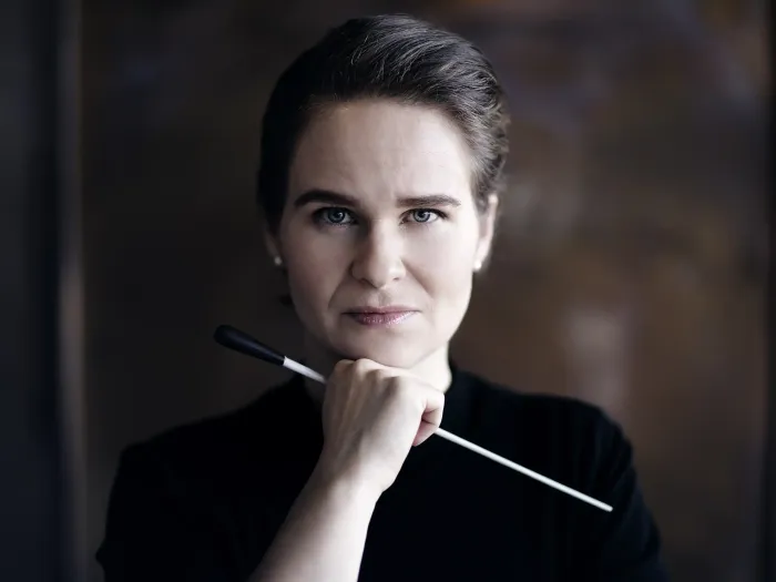 Person mit zurückgebundenem Haar hält einen Dirigentenstab vor dem Kinn, trägt schwarzen Pullover