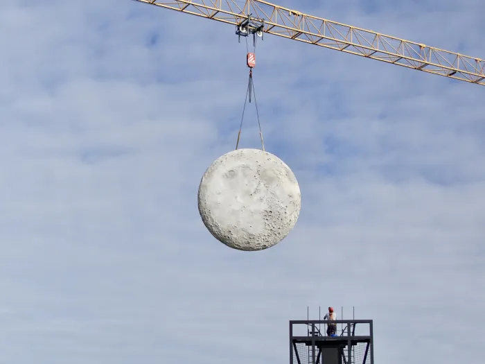 Runder, kraterübersäter Mond wird von einem Baukran an zwei Seilen über einem Gerüst mit einer Person gehoben.