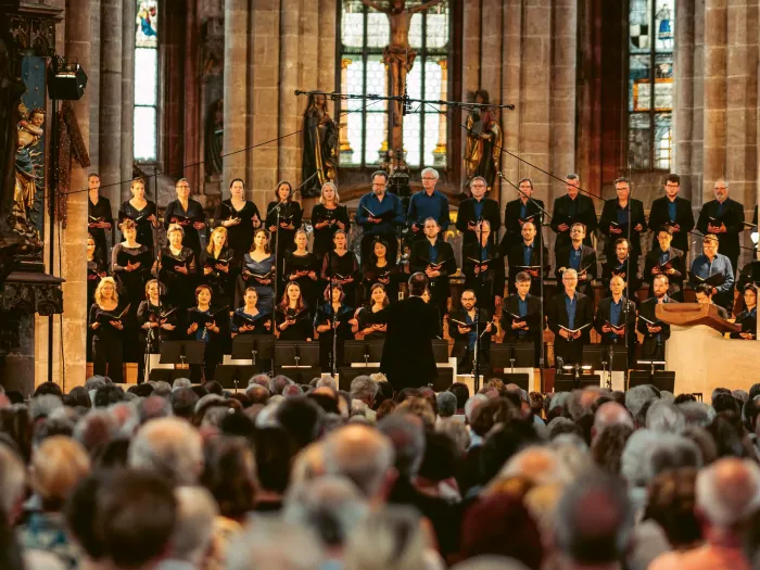 Großer Chor mit mehreren Sängerinnen und Sängern in dunkler Kleidung steht auf Stufen in einer Kirche vor Publikum, Dirigent mit erhobenen Armen.
